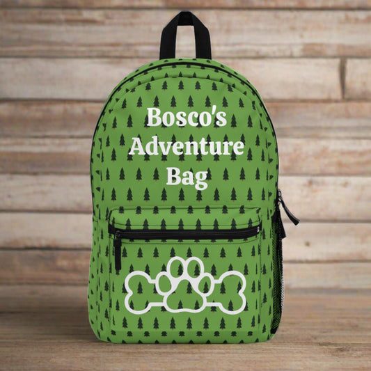 Front of green backpack with pine tree pattern. The top front has "Bosco's Adventures Bag" and the front of the pocket featuring an outline of a dog bone with a paw print in the middle of it. 