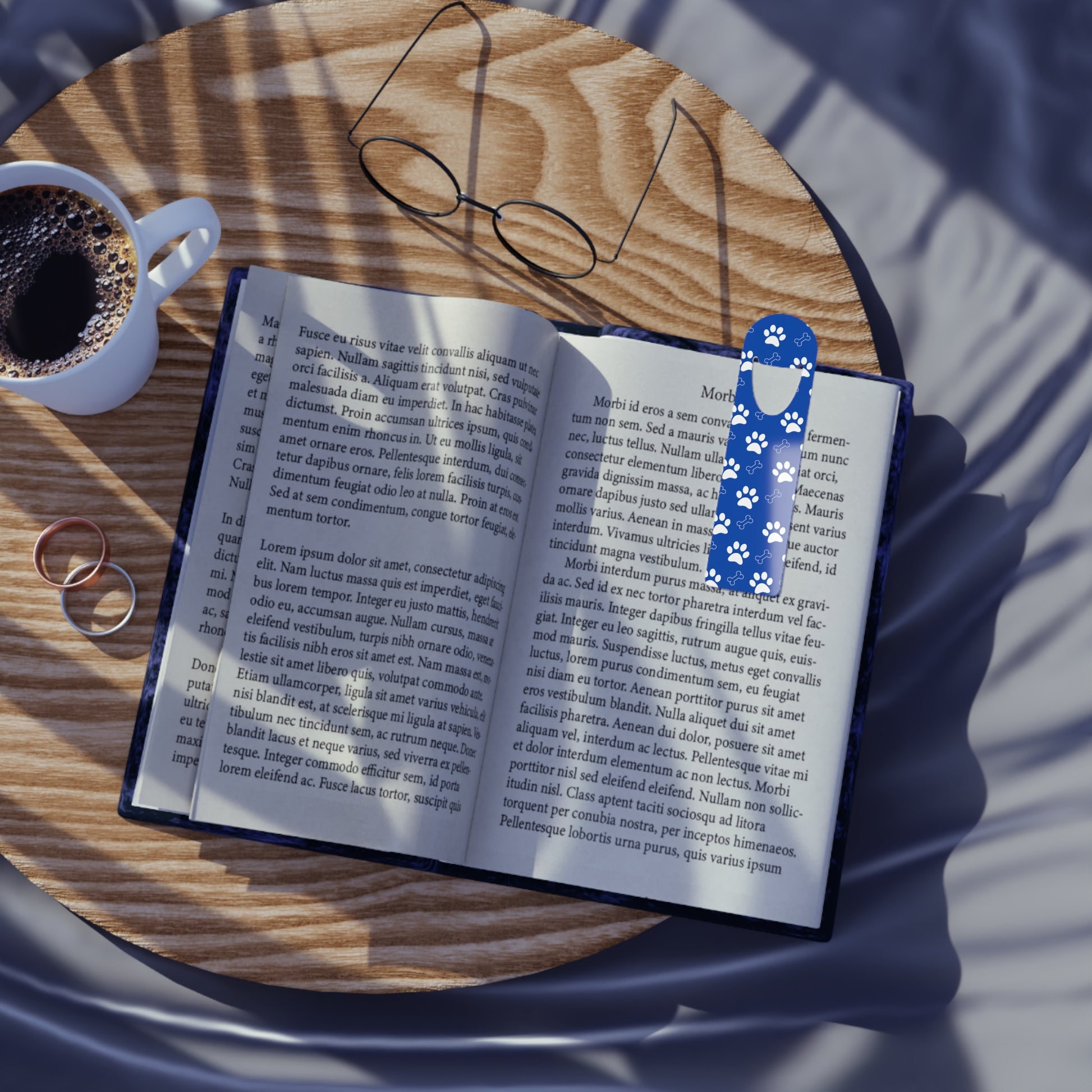 Blue Dog Paw Print Aluminum Book Mark in a book on a table with two wedding rings, a cup of black coffee and a pair of round spectacle glasses